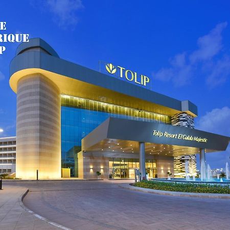 Tolip Resort El Galala Majestic Ain Sukhna Exterior foto The photo shows a modern hotel building named "Tolip Resort & Spa," featuring a sleek architectural design with large glass windows. The hotel is illuminated against a twilight sky, showcasing its prominent signage. In the foreground, there is a well