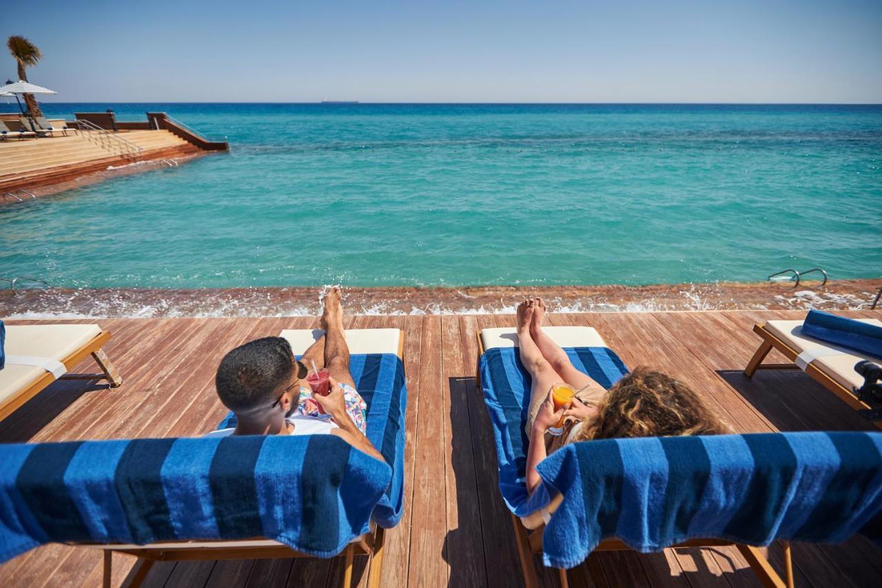 Tolip Resort El Galala Majestic Ain Sukhna Exterior foto The photo depicts two people relaxing on lounge chairs by the waterfront. They are positioned on a wooden deck with a view of the clear, turquoise water. Both individuals are reclining, enjoying the sun, and each has a drink in hand. The scene convey