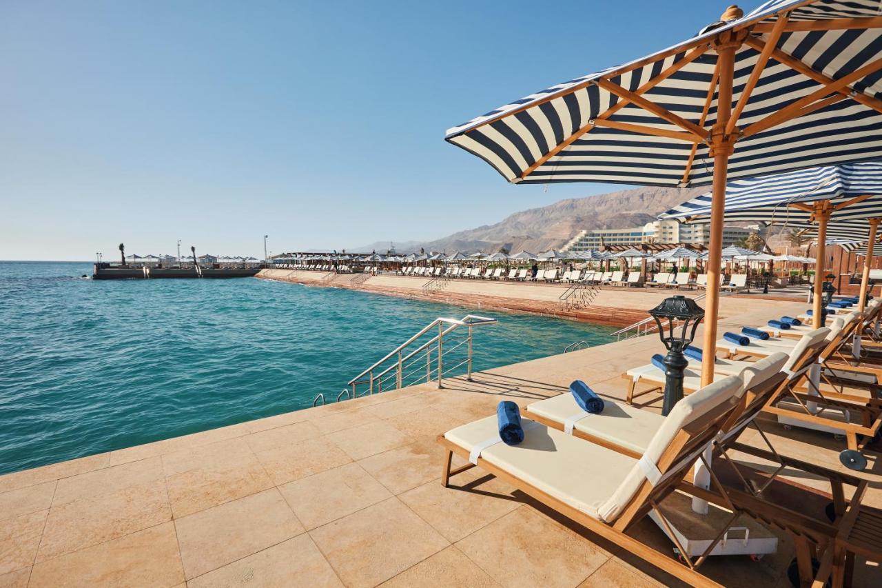 Tolip Resort El Galala Majestic Ain Sukhna Exterior foto The photo shows a scenic beachside area with clear blue water. In the foreground, there are several sun loungers set up on a tiled surface, with blue towels neatly rolled on each one. The loungers are shaded by striped umbrellas. In the distance, a w