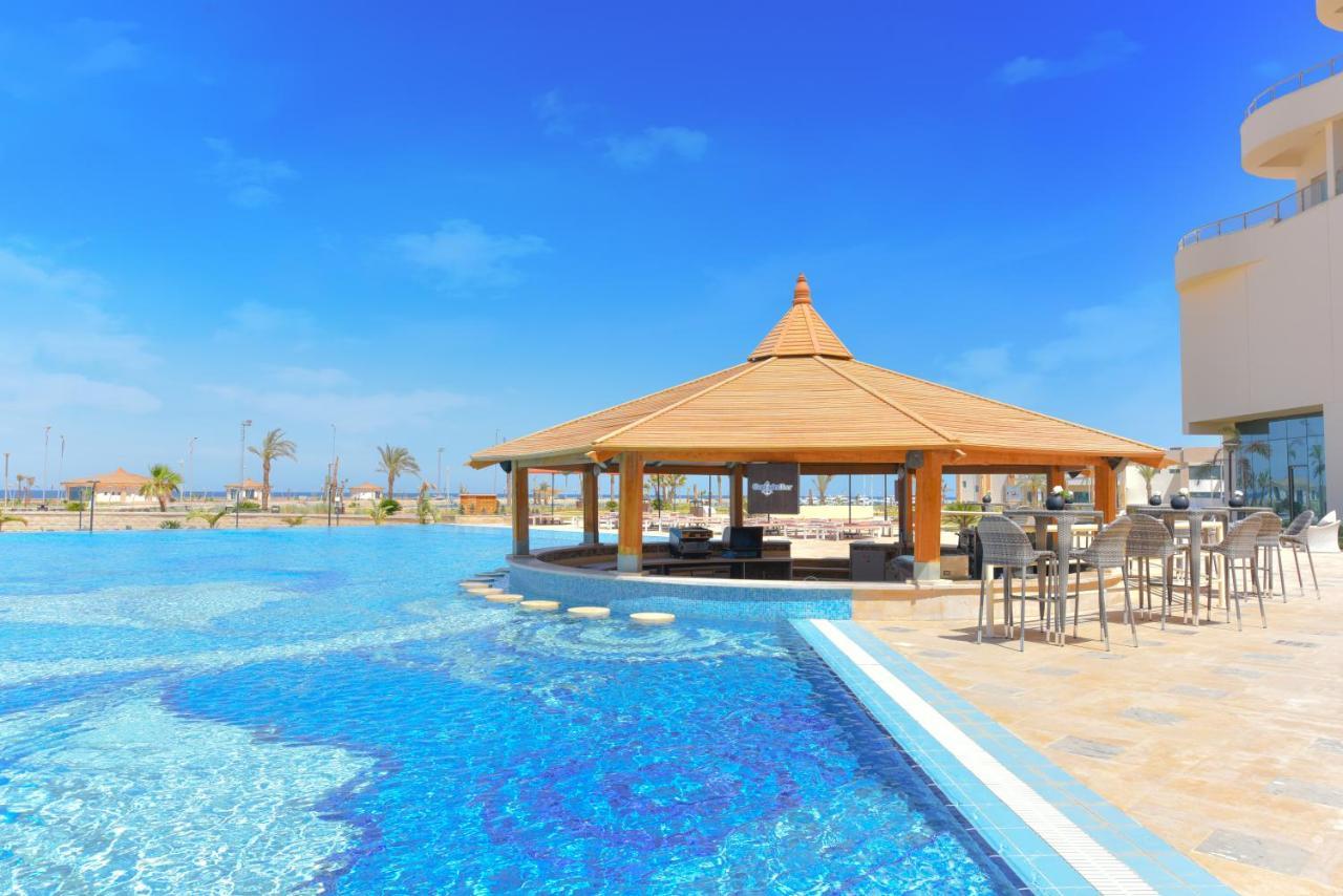 Tolip Resort El Galala Majestic Ain Sukhna Exterior foto The photo shows a beautiful outdoor setting featuring a large swimming pool with clear blue water. In the center, there is a thatched-roof gazebo, which serves as a shaded seating area. Surrounding the gazebo are lounge chairs and a bar area with hig