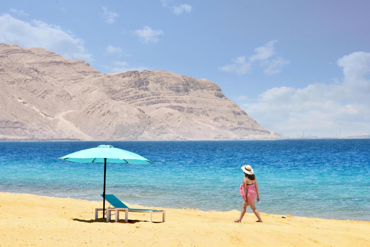 Tolip Resort El Galala Majestic Ain Sukhna Exterior foto The photo depicts a serene beach scene. In the foreground, there is a sandy shore with a single beach chair and an umbrella in a light blue color. A person dressed in a pink outfit is walking along the shoreline, enjoying the beach atmosphere. In the